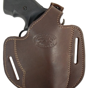 Op de afbeelding: Een bruine leren holster met een gestikt ontwerp en een riemlus. De holster heeft een uitsparing voor een pistool en de merknaam "Frontier Leather Company" is gegraveerd op de voorkant.