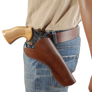 May include: A brown leather holster with a stitched edge is worn on a person's right hip. A wooden-handled revolver is inside the holster. The person is wearing a light brown shirt and blue jeans.