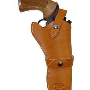 Może przedstawiać: Brązowa skórzana kabura z czarnym pistoletem i drewnianym uchwytem. Kabura ma pasek z metalowym zatrzaskiem i białymi szwami. Widoczna jest nazwa marki "CARBON LEATHER HANDCRAFTED".