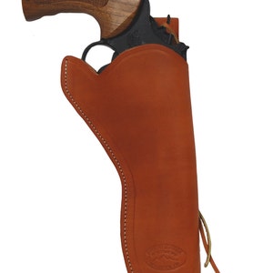 Op de afbeelding: Een zwarte revolver met een houten handvat zit gedeeltelijk in een bruinleren holster. De holster is gestikt met witte draad en heeft een logo. Een leren veter zet de holster vast. De holster is ongeveer 25 cm lang.