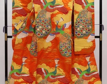 May include: An orange kimono with a vibrant peacock design. The kimono features embroidered peacocks with green bodies, red wings, and colorful tail feathers. Gold clouds and accents add to the ornate design. The garment is displayed on a black stand.