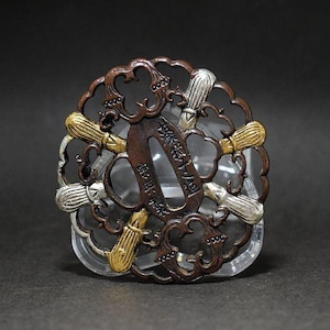 Puede incluir: Una tsuba circular de metal marrón oscuro con un intrincado diseño calado y elementos decorativos alternos en tonos oro y plata que parecen pinceles. La abertura ovalada central contiene caracteres japoneses grabados en el metal.