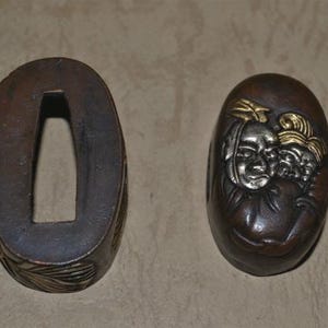 May include: Two antique Japanese tsuba, or sword guards, displayed side-by-side. One is a dark brown oval with a rectangular opening. The other is a dark brown oval with a detailed relief of faces and gold accents.