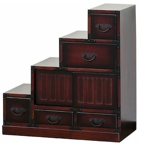 May include: A dark brown wooden step chest with multiple drawers and a cabinet. The chest has a stepped design, with each level featuring a drawer or cabinet. The drawers have ornate, dark metal handles and are framed with dark metal corner accents.