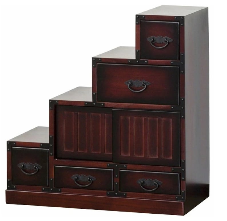 May include: A dark brown wooden step chest with multiple drawers and a cabinet. The chest has a stepped design, with each level featuring a drawer or cabinet. The drawers have ornate, dark metal handles and are framed with dark metal corner accents.