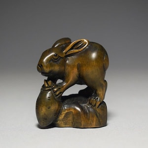 May include: A carved wooden sculpture of a rabbit perched atop a large root vegetable. The rabbit is brown with detailed features, including its ears and paws. The sculpture has a smooth, polished finish and is set against a neutral background.