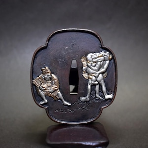 Puede incluir: Una guarda de espada de metal en tono bronce oscuro, o tsuba, con una abertura central cónica. La superficie muestra figuras en relieve de color plata y oro, incluyendo un individuo sentado sosteniendo un abanico y dos figuras luchando.