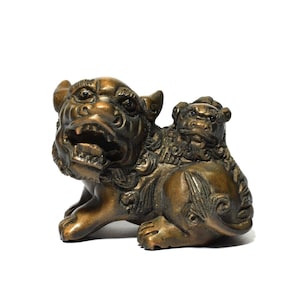 Puede incluir: Una figura de metal color bronce que representa un león guardián con la boca abierta, con un cachorro de león más pequeño descansando sobre su espalda. La escultura tiene un acabado metálico liso con tallas detalladas en caras, melenas y patas.