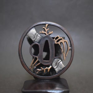Op de afbeelding: Een close-up van een Japanse tsuba, een zwaardbeschermer. De cirkelvormige tsuba heeft gedetailleerde uitgesneden ontwerpen en zilveren en gouden accenten. Het metaal heeft een donkere, verouderde uitstraling, wat suggereert dat het antiek is.