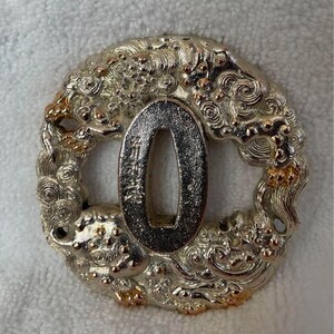 Op de afbeelding: Een tsuba, een decoratieve zwaardbeschermer, van zilver en goud, met een gedetailleerd ontwerp van een draak in wolken. De tsuba is rond en heeft een gat in het midden voor het zwaardblad.