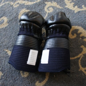 Puede incluir: Un par de guantes de kendo acolchados negros y azules con parches blancos. Los guantes están sobre una alfombra estampada.
