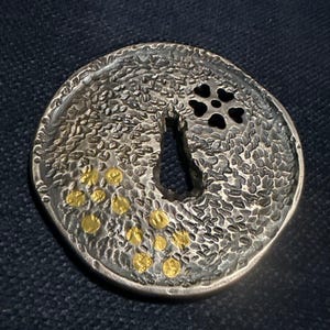 May include: A round, silver-toned metal object with a textured surface and a keyhole-shaped opening. The design includes a four-petal flower shape and several gold-colored dots. The object has a rustic, handcrafted appearance.