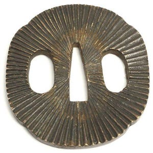 Op de afbeelding: Een bronskleurige, ovale tsuba, een Japanse zwaardbeschermer. De tsuba heeft een centrale opening en twee symmetrische openingen. Het oppervlak is getextureerd met stralende lijnen, waardoor een zonnestraaleffect ontstaat. Het ontwerp is gedetailleerd en ingewikkeld.