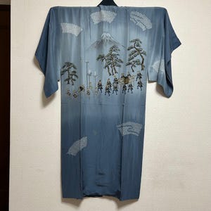 May include: A blue kimono with an embroidered landscape design. The design includes a mountain, trees, and figures in a procession. The kimono's colour scheme gradients from a lighter blue at the top to a darker blue at the bottom.