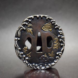 Op de afbeelding: Een close-up van een Japanse tsuba, een zwaardbeschermer. De cirkelvormige beschermer heeft een donkerbruine basis met gouden en zilveren bloempatronen. Het ontwerp omvat een chrysant en vloeiend gebladerte.