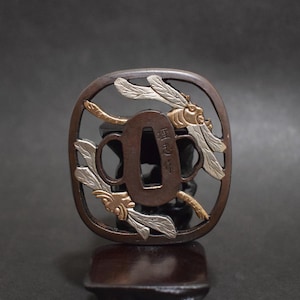 Op de afbeelding: Een bronskleurige Japanse tsuba, een zwaardbeschermer, met een libellenontwerp. De libellenvleugels zijn zilver en goud, en de tsuba heeft een rechthoekige opening in het midden. Japanse karakters zijn zichtbaar.