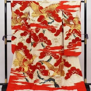 May include: A traditional Japanese kimono, primarily cream-colored, adorned with vibrant red and gold phoenix and cloud motifs. The design includes stylized trees and flowing red lines, creating a dynamic and elegant aesthetic. The kimono is displayed on a black stand.