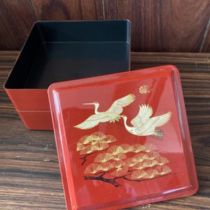 Może przedstawiać: Czerwona lakierowana skrzynia do układania w stos z czarnym wnętrzem i złotym wzorem dwóch żurawi lecących nad sosną.