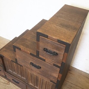 May include: A wooden step-style cabinet with multiple drawers. The cabinet is made of dark brown wood and has black metal accents. The drawers have black metal handles.