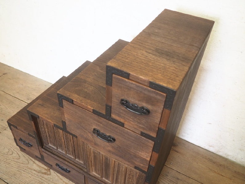 May include: A wooden step-style cabinet with multiple drawers. The cabinet is made of dark brown wood and has black metal accents. The drawers have black metal handles.