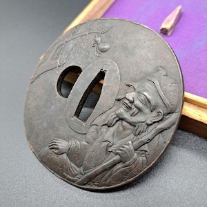 Op de afbeelding: Een ronde, bronzen tsuba, een decoratieve zwaardbeschermer, met een gedetailleerde reliëfgravure van een lachende man die een staf vasthoudt. De man draagt een hoed en traditionele kleding. Een kleine spin is in het bovenste deel van de tsuba gegraveerd.
