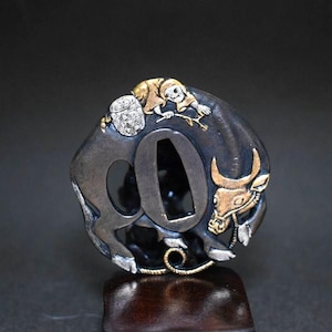 Op de afbeelding: Een ronde tsuba van donker metaal met een goudkleurige figuur op een stier. De stier heeft goudkleurige hoorns en een gezicht, terwijl de ruiter een dunne tak vasthoudt. Het object is voorzien van een centrale ovale opening en metaalwerk.