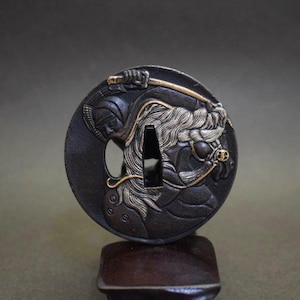 Op de afbeelding: Een gedetailleerde, zwart-gouden Japanse tsuba, een zwaardbeschermer. De ronde beschermer toont een krijgersfiguur met een zwaard en een paard met een vloeiende manen. Het ontwerp is ingewikkeld gesneden en gedetailleerd, met een donkere achtergrond en gouden accenten.