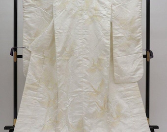 May include: A white kimono with a gold crane pattern is displayed on a black stand. The kimono is made of a textured fabric and has long sleeves. The crane design is repeated across the garment.