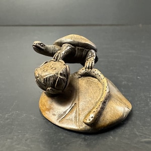 Op de afbeelding: Een klein bronzen beeldje van een schildpad die op een lotusblad zit. De schildpad is gedetailleerd met een getextureerde schild en een lange, gekrulde staart. Het lotusblad is ook getextureerd en heeft een verhoogd midden.