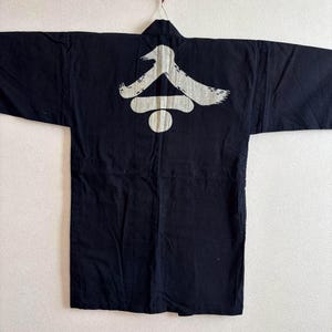 Puede incluir: Chaqueta negra estilo kimono con un gráfico blanco en la espalda. El gráfico parece ser un símbolo japonés estilizado. La chaqueta está colgada de una percha sobre un fondo neutro. Las mangas son anchas y la chaqueta está abierta por delante.