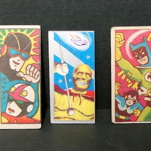 May include: Three vintage trading cards featuring illustrations of superheroes. The first card features a masked superhero in a yellow and black costume. The second card features a superhero with a skull mask holding a staff. The third card features a superhero in a green and yellow costume with a bat symbol on the chest.