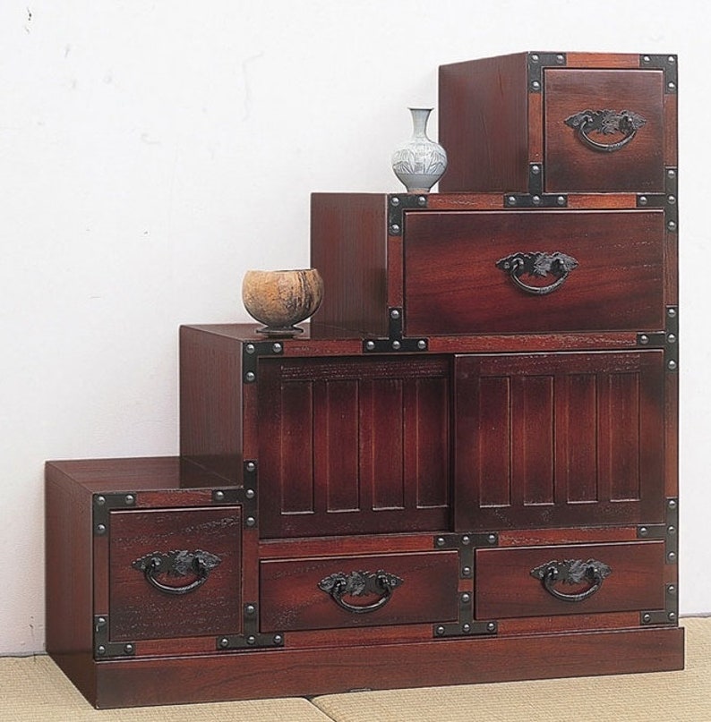May include: A dark reddish-brown wooden step chest with multiple drawers and compartments. The chest has decorative metal corner accents and ornate black handles. A small vase and a bowl sit on top of the chest.