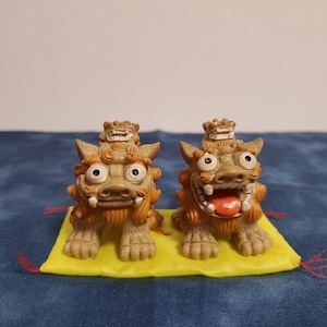 May include: Two tan and orange Shisa statues, traditional Ryukyu guardian lions, sit on a yellow mat. One has an open mouth with a red tongue, the other is closed. Each has a smaller Shisa on its head.