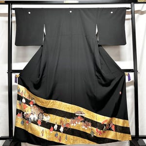 May include: A black kimono with a decorative gold and black striped border featuring a detailed illustration of people and carriages. The kimono is displayed on a black metal stand, showcasing its traditional design and craftsmanship.
