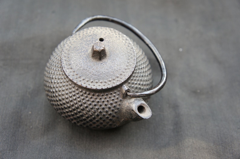 Op de afbeelding: Een zwarte gietijzeren theepot met een getextureerd, gebollet oppervlak en een kleine tuit. De theepot heeft een deksel en een handvat.