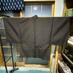 May include: A dark brown traditional Japanese kimono hanging on a wooden rack. The kimono is made of a smooth fabric and has wide sleeves.