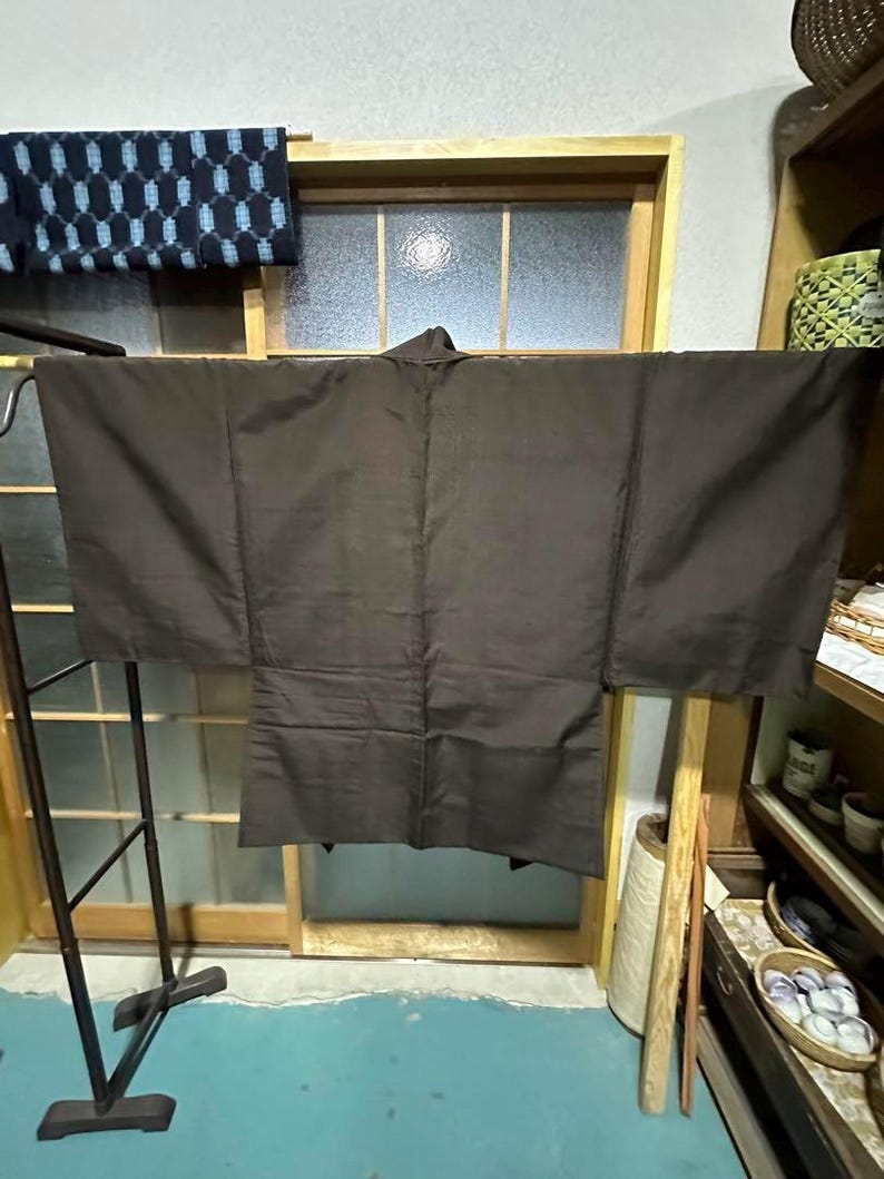 May include: A dark brown traditional Japanese kimono hanging on a wooden rack. The kimono is made of a smooth fabric and has wide sleeves.