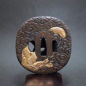 Puede incluir: Una guarda de espada circular de bronce oscuro, conocida como tsuba, con un patrón en relieve de nubes arremolinadas. Un dragón de color dorado aparece emergiendo de las nubes en la parte inferior izquierda, con una pequeña forma de luna creciente dorada arriba a la derecha.