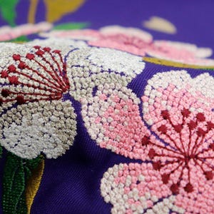 May include: Close-up of embroidered fabric with pink, white, and red flowers on a purple background. The flowers are detailed and intricate, with small stitches creating a textured effect.