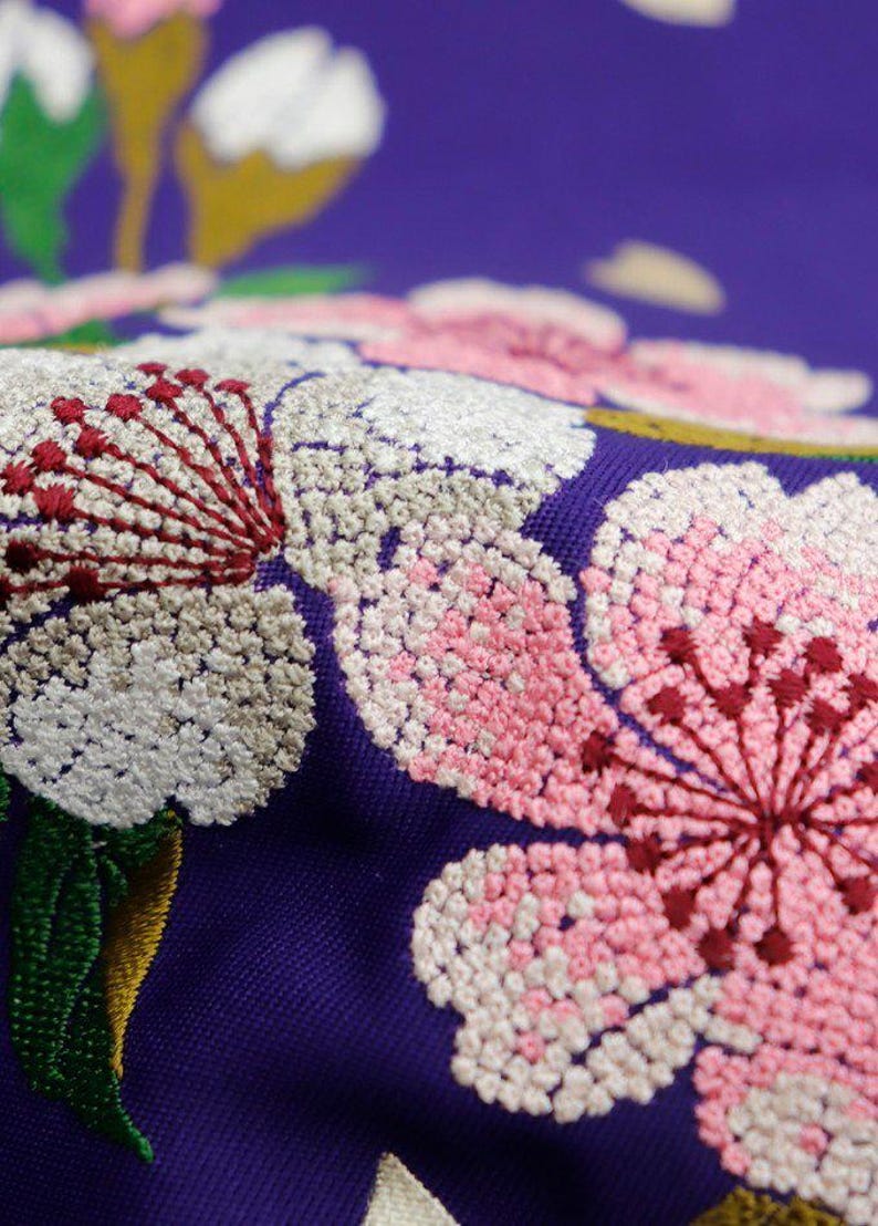 May include: Close-up of embroidered fabric with pink, white, and red flowers on a purple background. The flowers are detailed and intricate, with small stitches creating a textured effect.