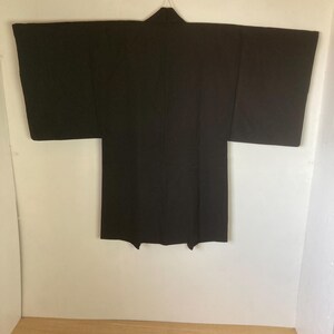 May include: A black Japanese traditional garment, a haori, hangs on a hanger against a white wall. The haori is a short, hip-length jacket worn over a kimono.