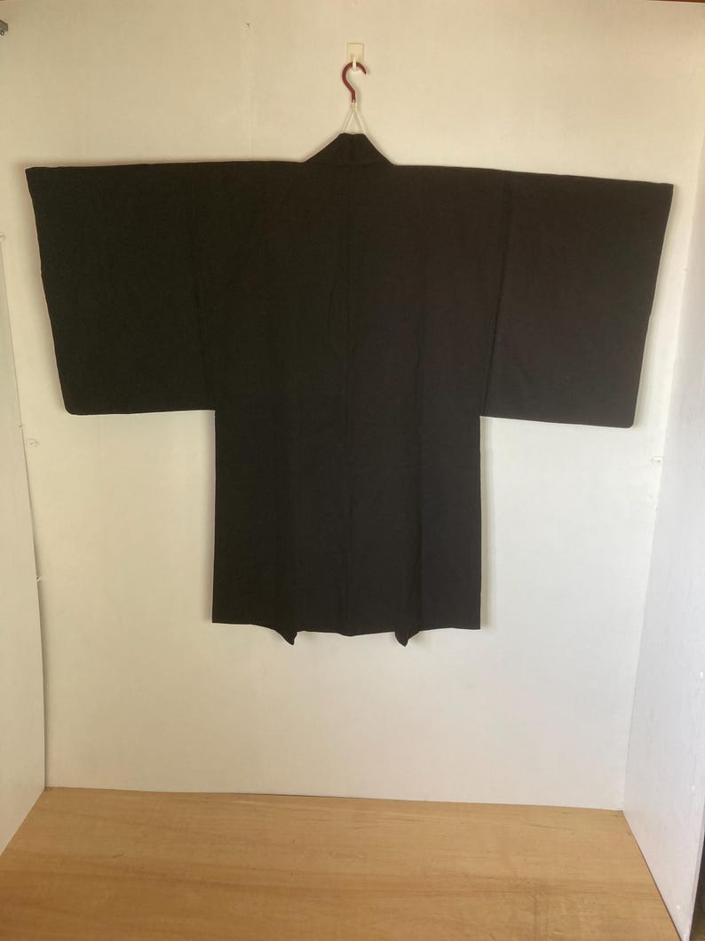 May include: A black Japanese traditional garment, a haori, hangs on a hanger against a white wall. The haori is a short, hip-length jacket worn over a kimono.