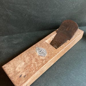 Puede incluir: Un cepillo de mano de madera vintage con una cuchilla de metal. El cepillo está hecho de madera de color claro y tiene un cuerpo rectangular. Una cuchilla de metal está colocada en la parte superior del cepillo. Un emblema decorativo está en el lateral del cepillo.