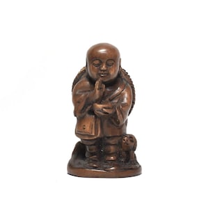 May include: A brown carved wooden figurine depicting a monk with a shaved head, wearing robes, and holding hands in a prayer position. A small dog stands at the monk's side, and a circular woven-style pack is visible on the monk's back.
