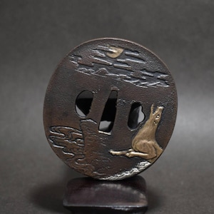 Op de afbeelding: Een ronde tsuba van donker metaal met een reliëfontwerp van een gouden wolf die naar de maan huilt boven gestileerde wolken en water. De zwaardstootplaat heeft een centrale opening en twee kleinere openingen, met een donkerbruine patina afwerking.