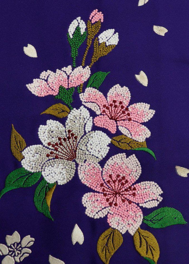 May include: Embroidered cherry blossoms in shades of pink, white, and green on a deep purple fabric background. The flowers are detailed with small beads and the leaves are a vibrant green.
