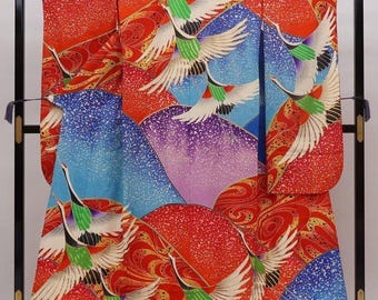 May include: A vibrant, patterned kimono featuring a design of flying cranes against a backdrop of stylized mountains. The color palette includes red, blue, purple, and white, with the cranes' wings spread wide. The garment is displayed on a black stand.