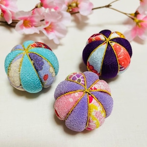 Puede incluir: Tres bolas decorativas japonesas Temari. Cada bola está hecha de paneles de tela en tonos de azul, morado, rosa y amarillo, cosidos con hilo dorado. Las bolas están dispuestas sobre una superficie blanca con ramas de flores de cerezo rosas en el fondo.