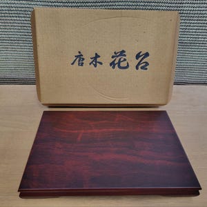 May include: A rectangular wooden display stand with a deep reddish-brown wood grain finish, positioned in front of a tan cardboard box featuring dark blue Chinese characters printed on the front surface of the packaging.