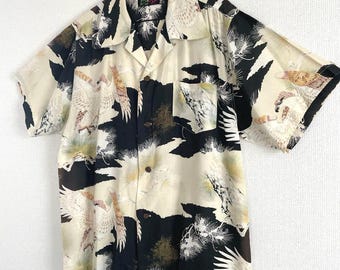 May include: Short-sleeved button-down shirt with a cream, black, and tan pattern. The shirt features a bird and foliage design, a chest pocket, and a classic collar. The shirt is hanging on a black hanger.
